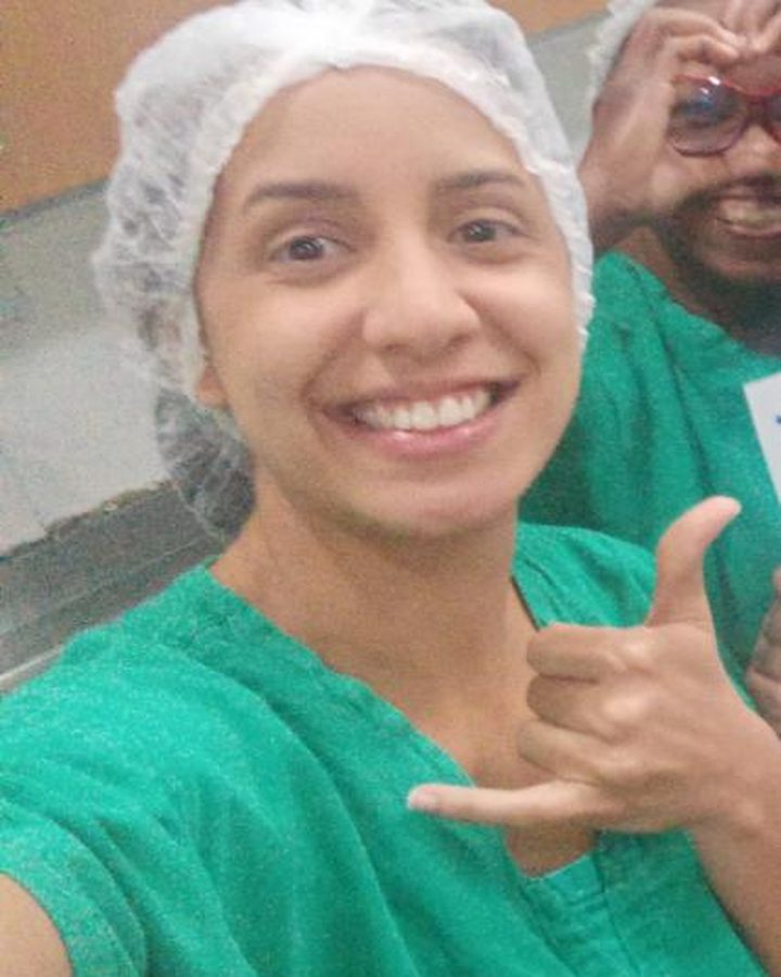 Camila Cardoso sorrindo de touca e pijama cirúrgico verde, ao lado de um colega de equipe no centro cirúrgico