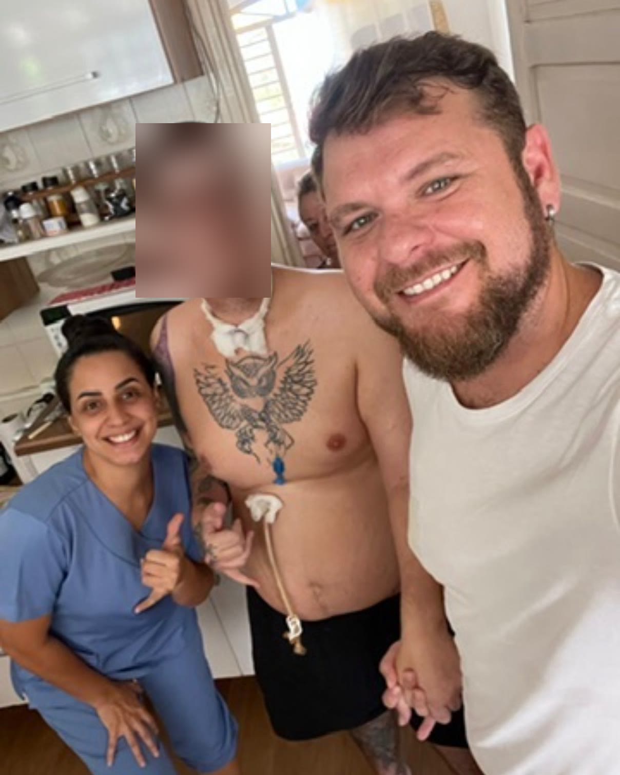 Camila Cardoso, de pijama cirúrgico, sorrindo ao lado do paciente que acompanhou no home care e de um familiar, dentro da casa da família; o rosto do paciente está borrado para preservar sua identidade