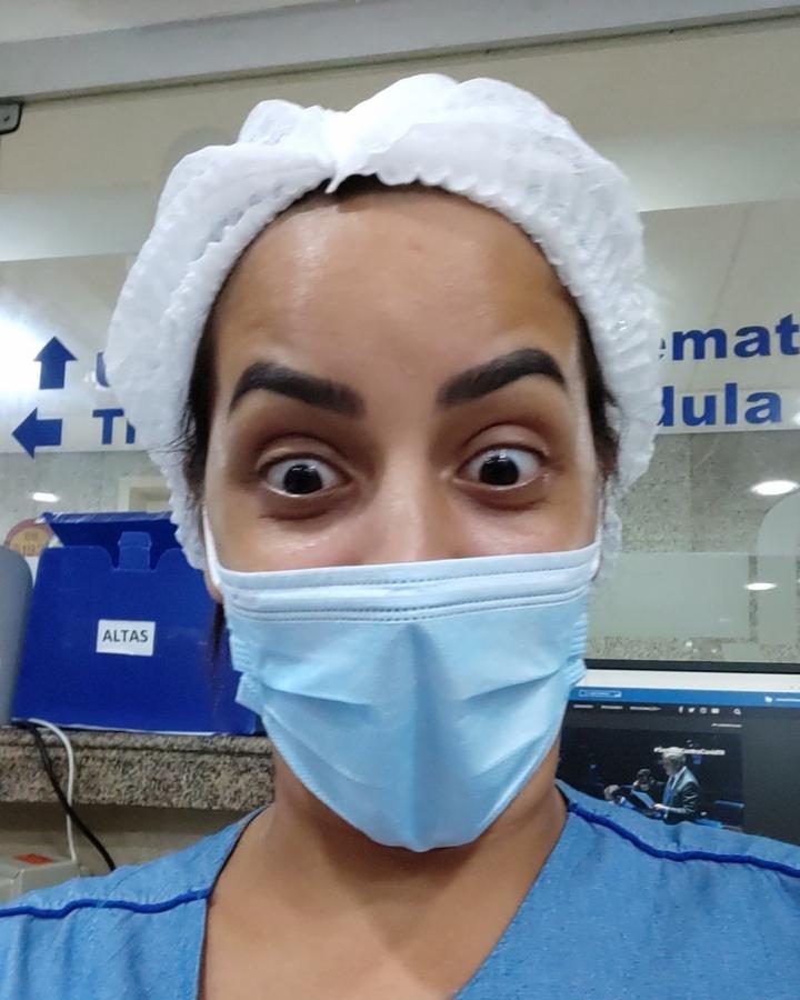 Camila Cardoso de touca e máscara cirúrgica, com os olhos arregalados, no balcão de altas do hospital