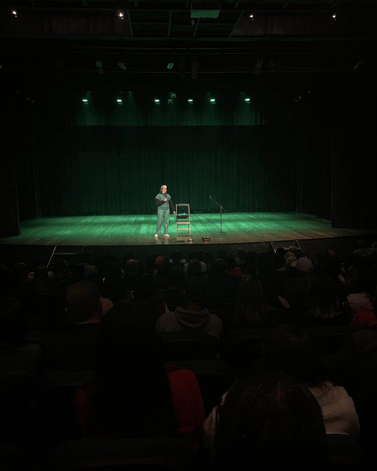 Camila Cardoso no palco, de microfone na mão, durante show de stand-up com plateia lotada