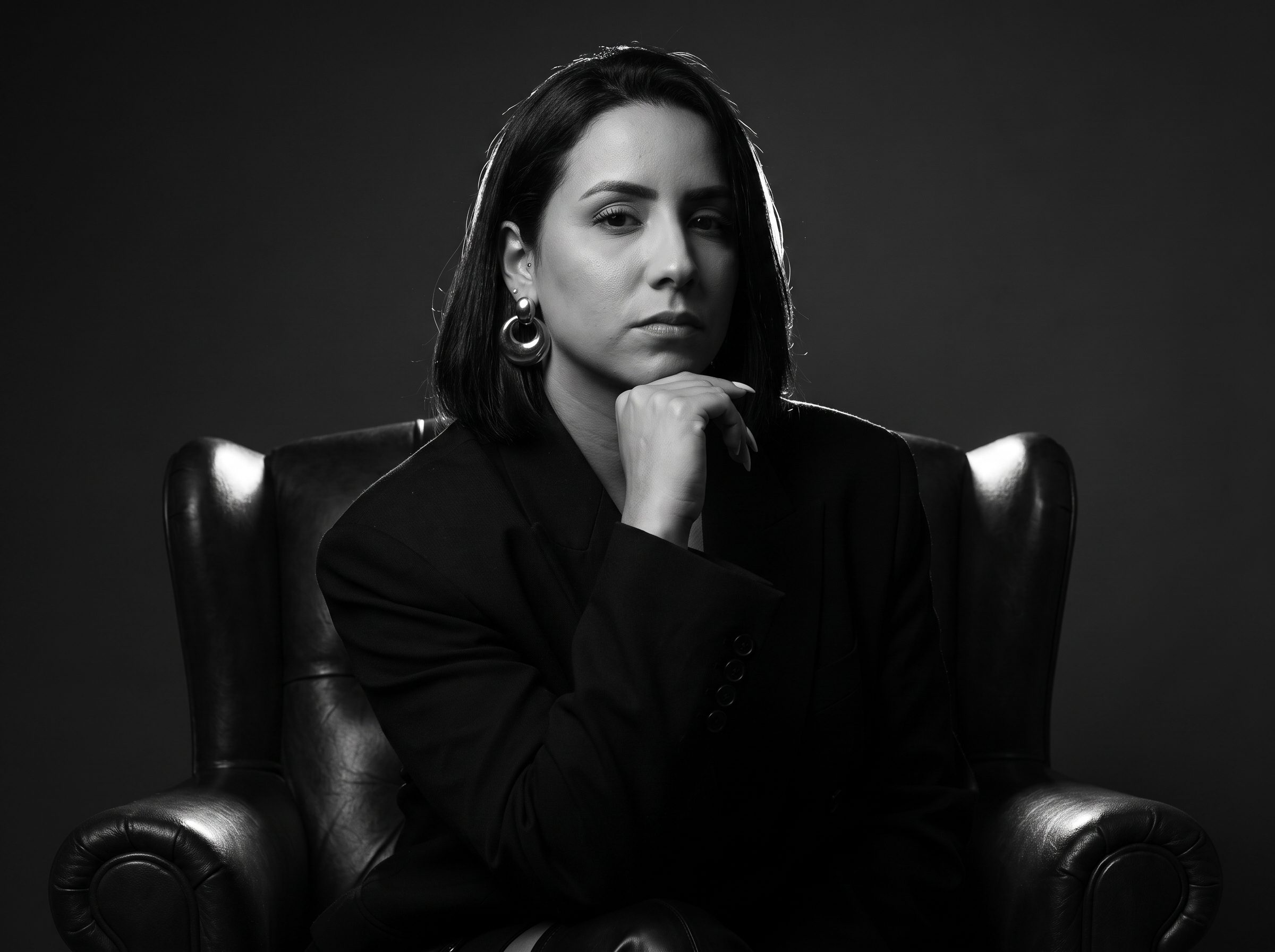 Camila Cardoso, de blazer, sentada em uma poltrona de couro em retrato preto e branco