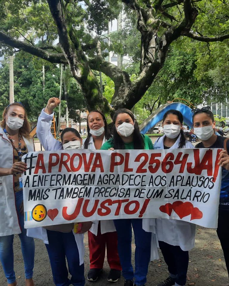 Profissionais de enfermagem de jaleco e máscara em manifestação, segurando uma faixa: “#Aprova PL 2564 já — a enfermagem agradece aos aplausos mas também precisa de um salário justo!”