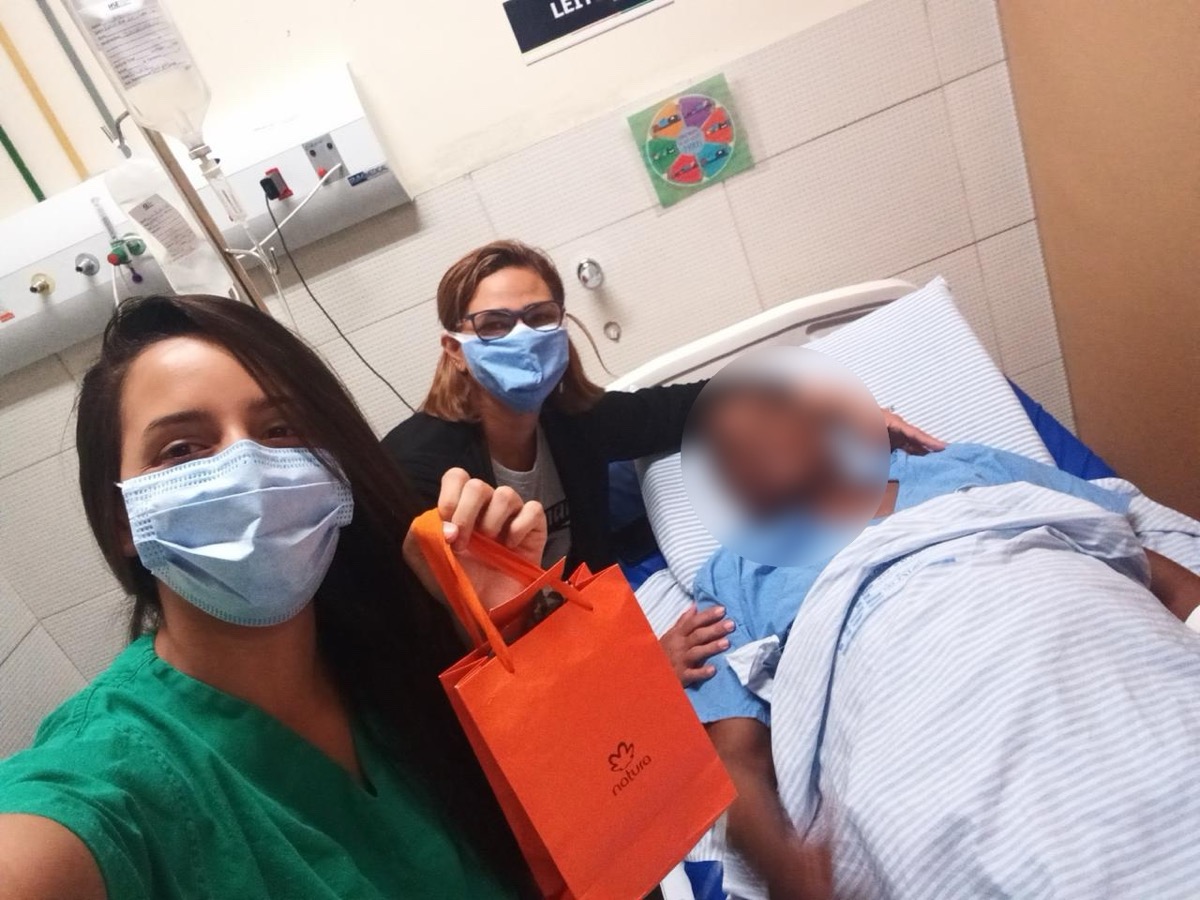 Camila Cardoso e colega entregam um presente a um paciente internado; o rosto do paciente está borrado para preservar sua identidade