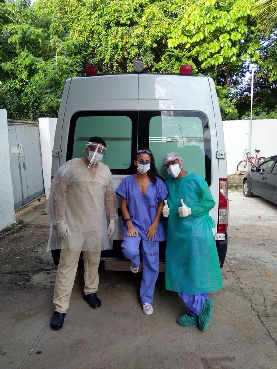 Camila Cardoso entre dois colegas paramentados atrás de uma ambulância